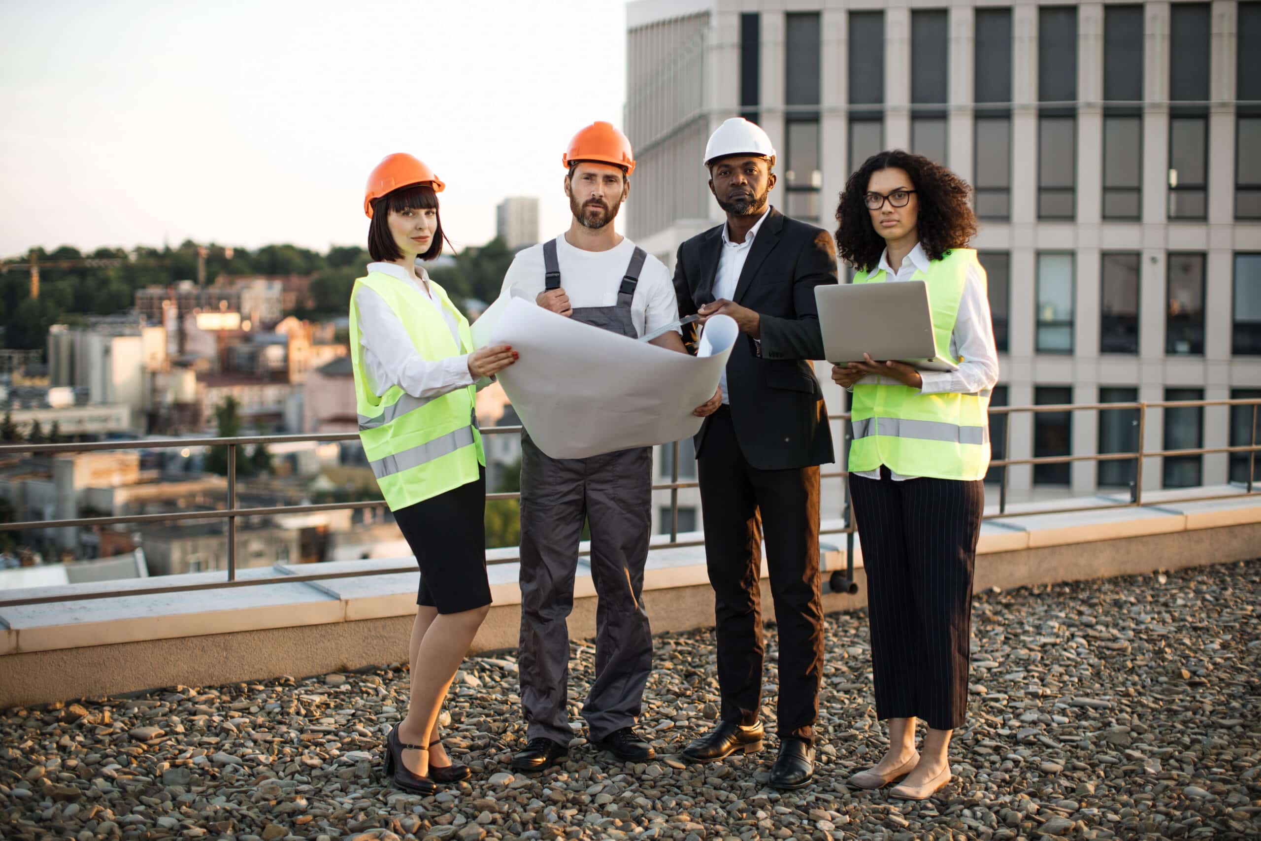 Full length portrait of multiethnic work team standing on terrace with amazing city view and looking at camera. Four international urban planners dressed in protective wear holding layouts and laptop.